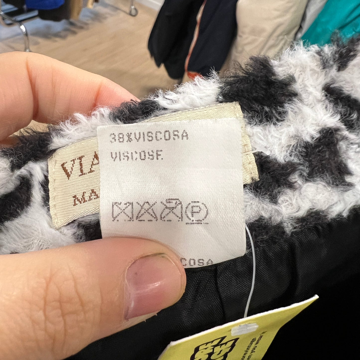 Close-up of a white care and composition label on a black-and-white textured garment, showing 38% viscose and washing symbols, held between two fingers