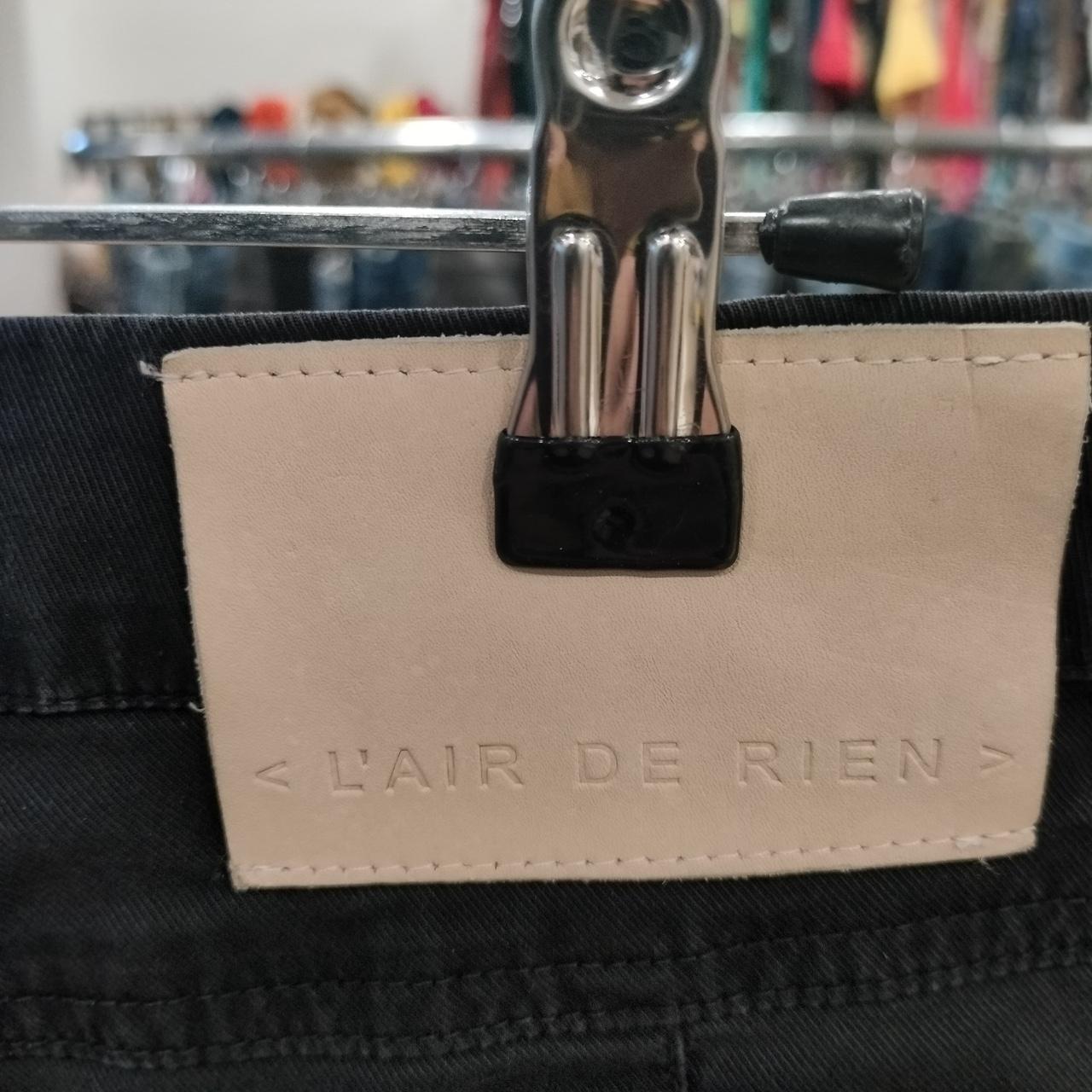 Beige leather label with 'L'AIR DE RIEN' on black denim, clothes rack background