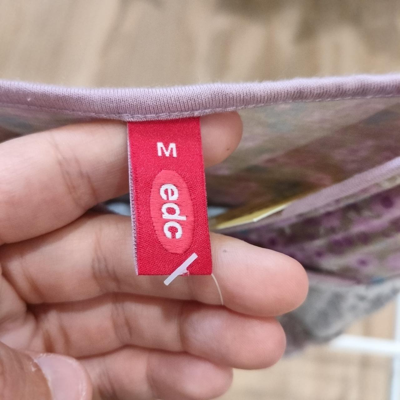 Hand holding red clothing tag with 'M edc' label on patterned fabric