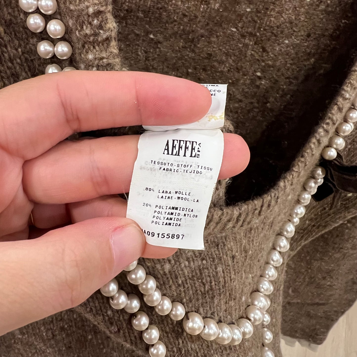 Hand holding a clothing label with 'AEFFES' branding against a textured fabric background.