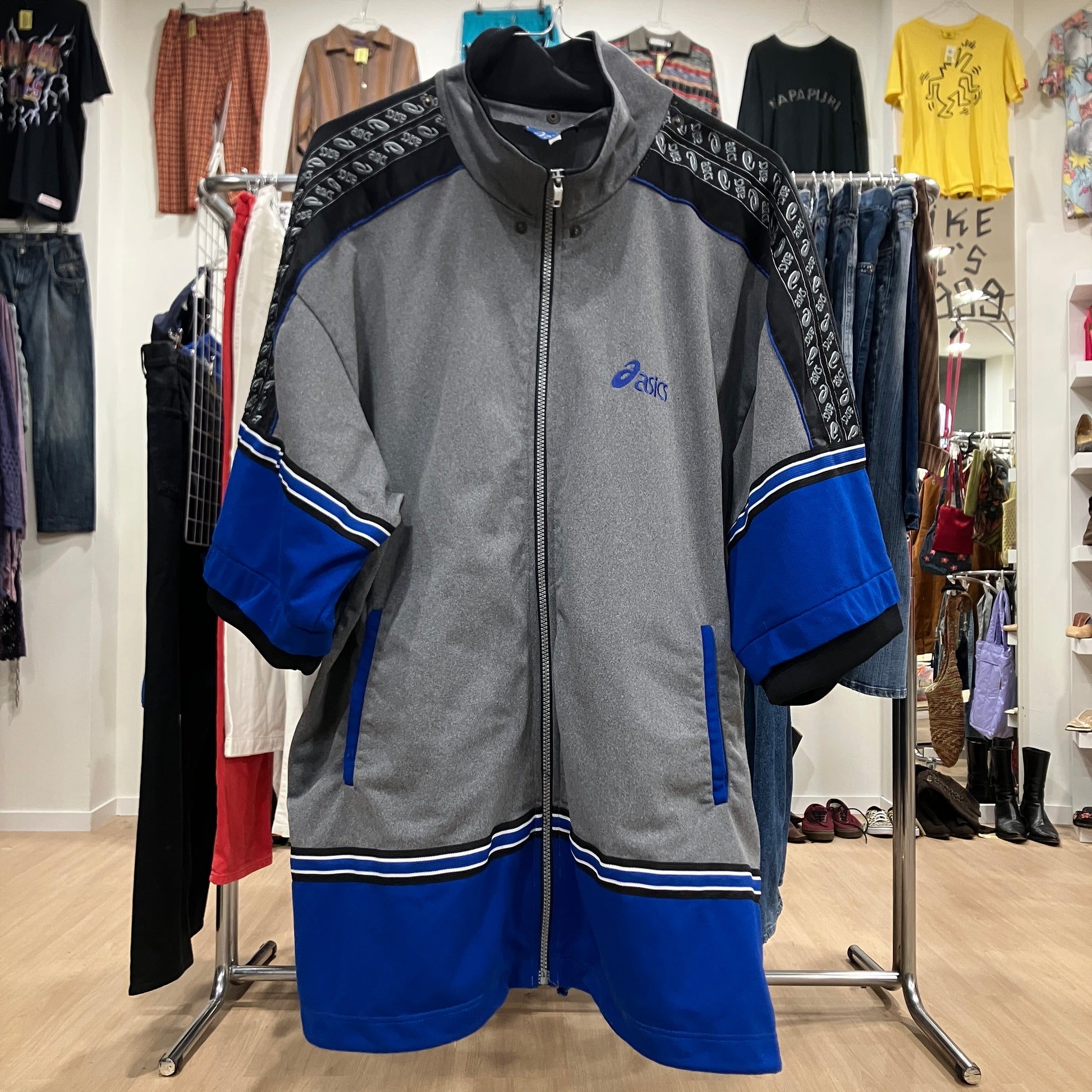 Front view of a vintage Asics zip-up jacket in grey, black, and blue with patterned shoulder panels and striped details on the sleeves and hem, displayed on a hanger inside a vintage clothing store

