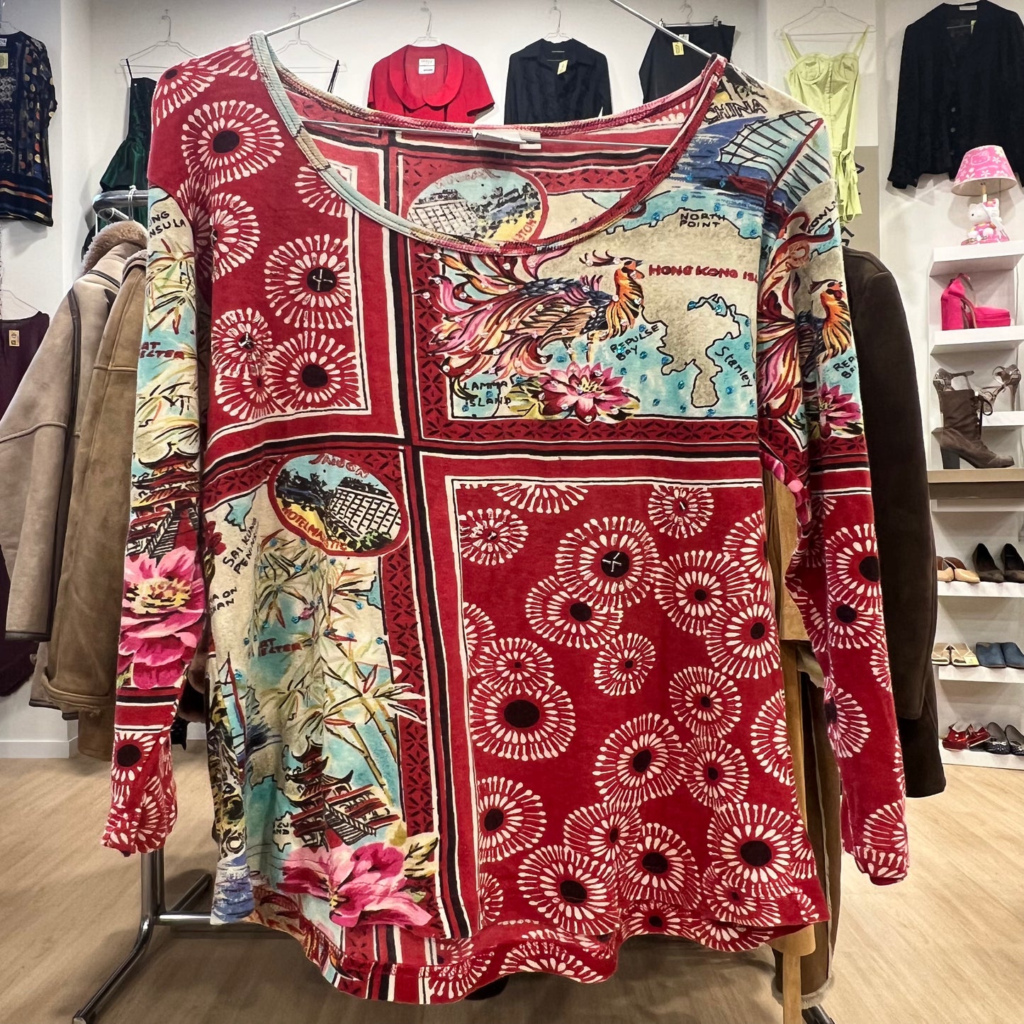 Long-sleeve bohemian top featuring a red base with a mixed travel and floral print, including maps, Asian motifs, and abstract circular patterns, displayed on a clothing rack in a store