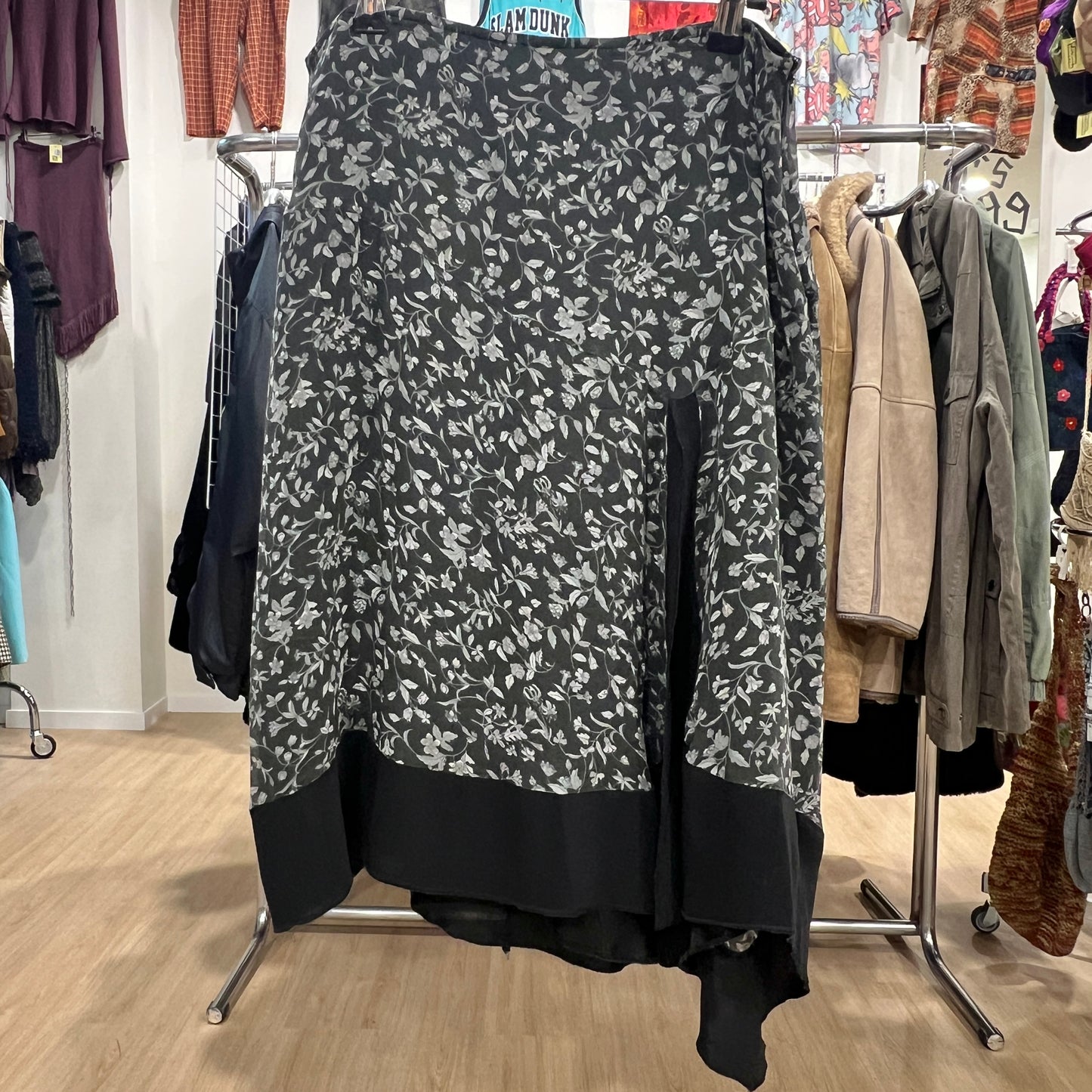 Front view of a black midi skirt with a grey floral pattern and a solid black asymmetrical hem, displayed on a hanger inside a vintage clothing store
