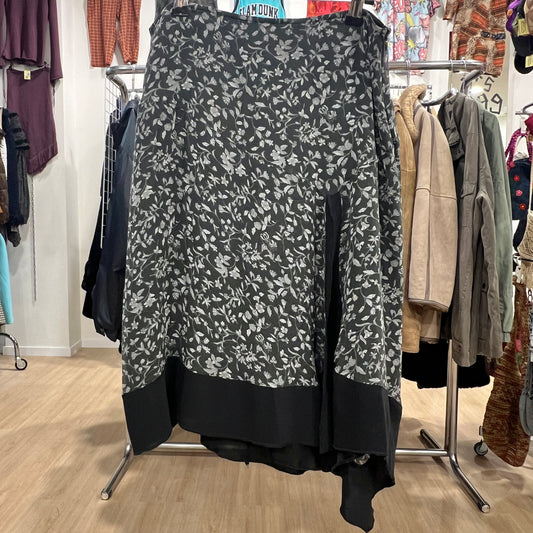 Front view of a black midi skirt with a grey floral pattern and a solid black asymmetrical hem, displayed on a hanger inside a vintage clothing store
