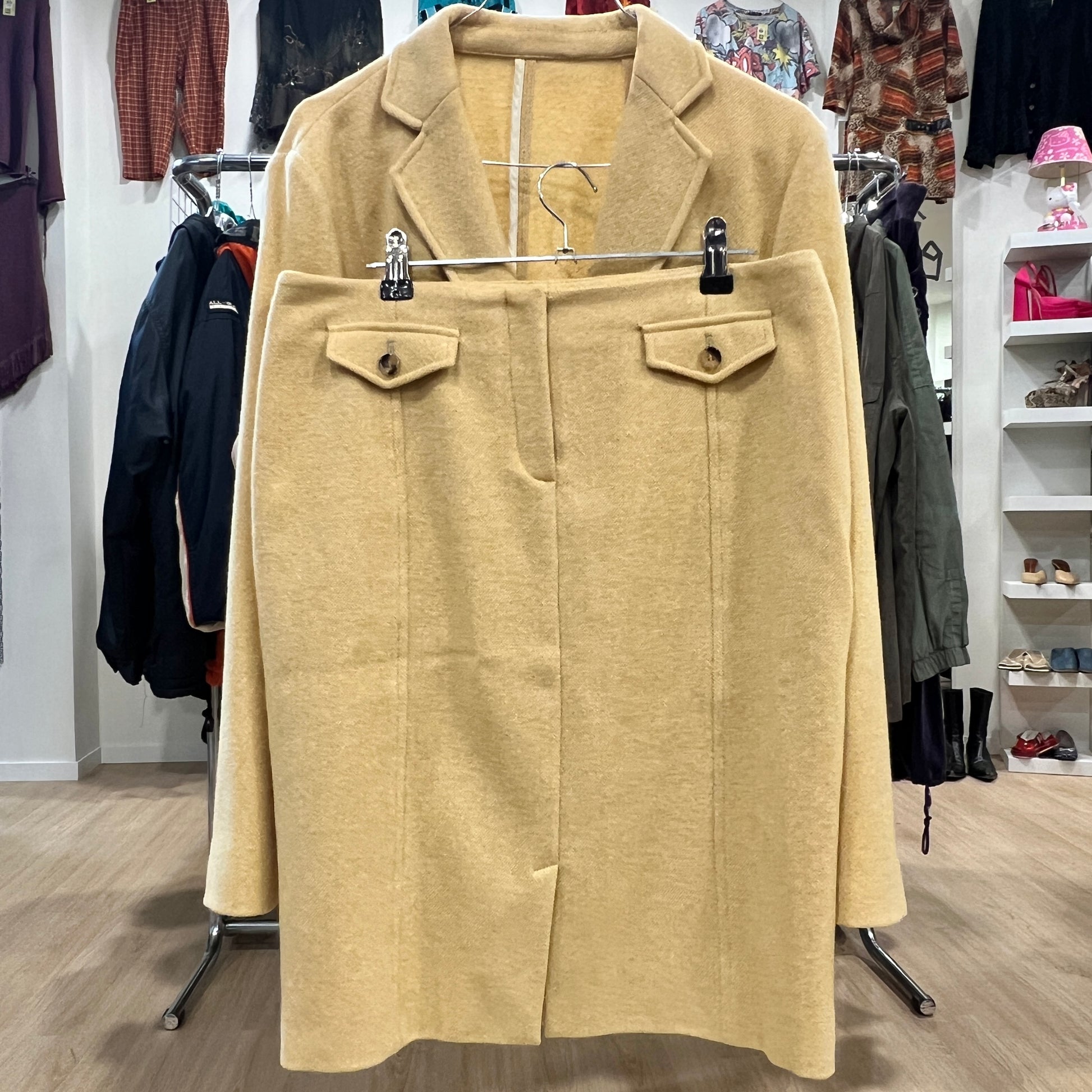 Front view of a beige wool skirt suit set with a knee-length skirt featuring buttoned flap pockets, displayed together with the matching blazer on a hanger inside a vintage clothing store