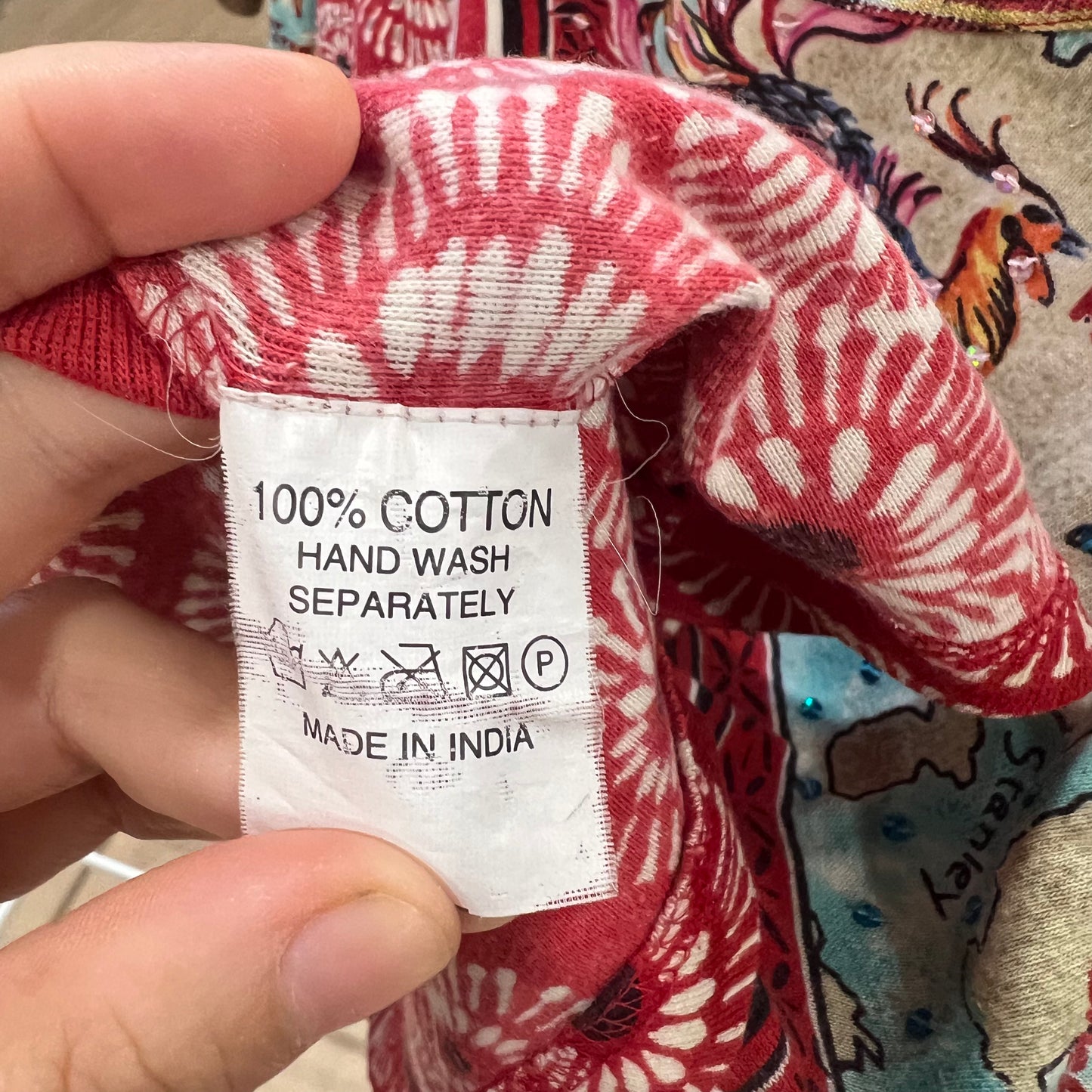 Hand holding a red and white patterned fabric with a label indicating 100% Cotton Hand Wash Separately Made in India
