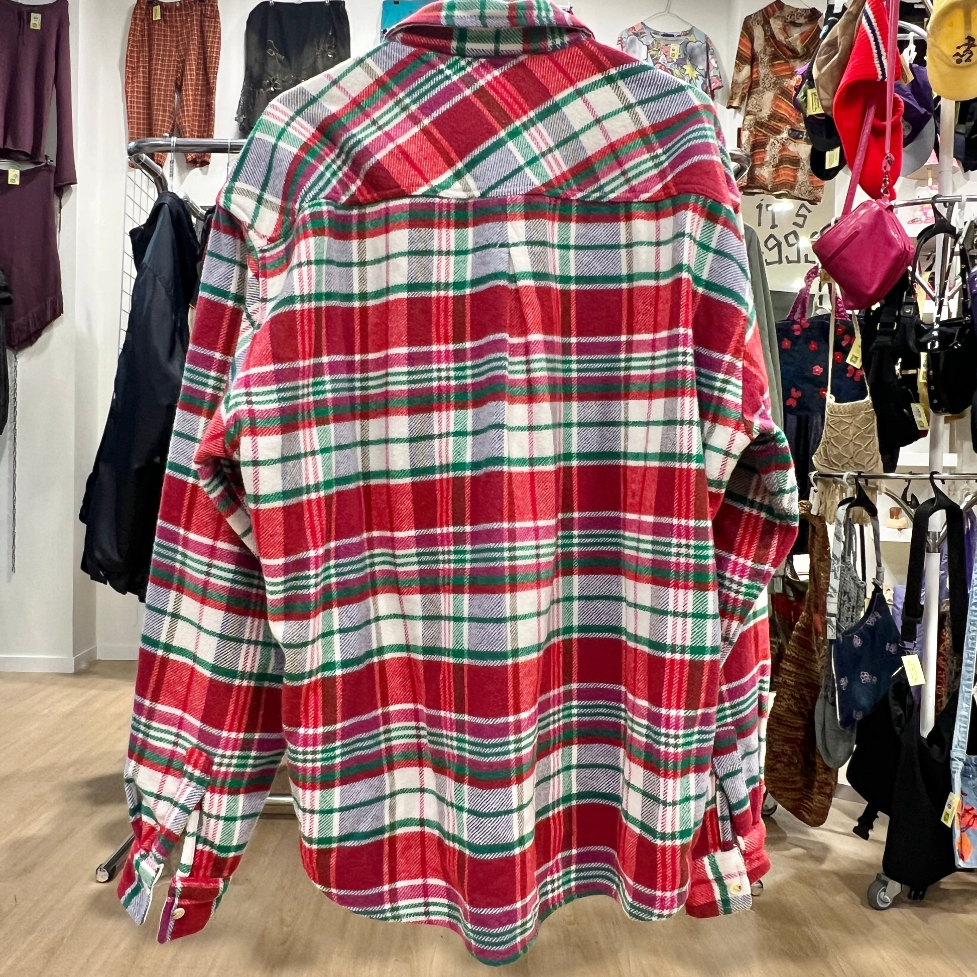 Red plaid shirt in a store setting with clothing racks and bags in the background