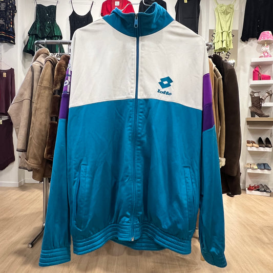 Vintage Lotto track jacket in teal, white, and purple color block design, featuring a zip-up front, high collar, and embroidered logo on the chest, displayed on a clothing rack in a store