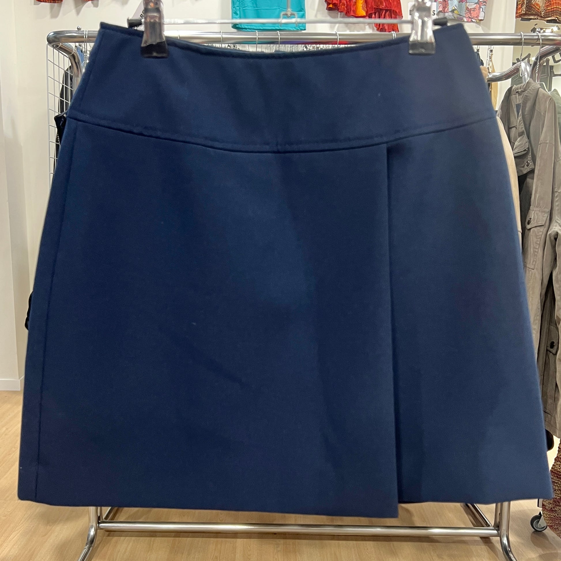 Front view of a navy blue A-line skirt with a clean structured design and a single front pleat, displayed on a hanger inside a vintage clothing store

