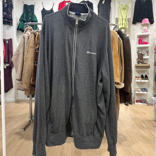 Dark gray Champion track jacket with a full zip closure, high collar, and small embroidered logo on the chest, displayed on a hanger in a clothing store