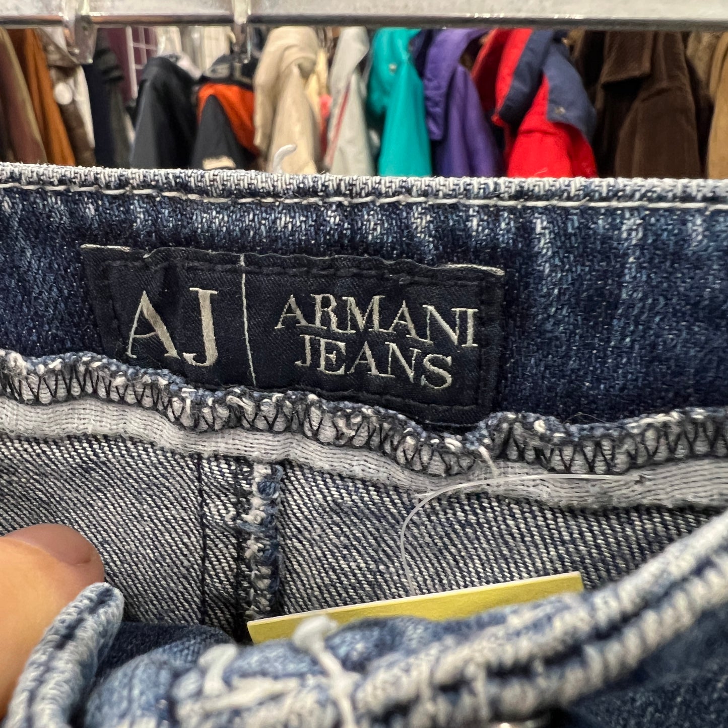 Close-up of a black AJ Armani Jeans label stitched onto the waistband of blue denim jeans, with visible contrast stitching and fabric texture
