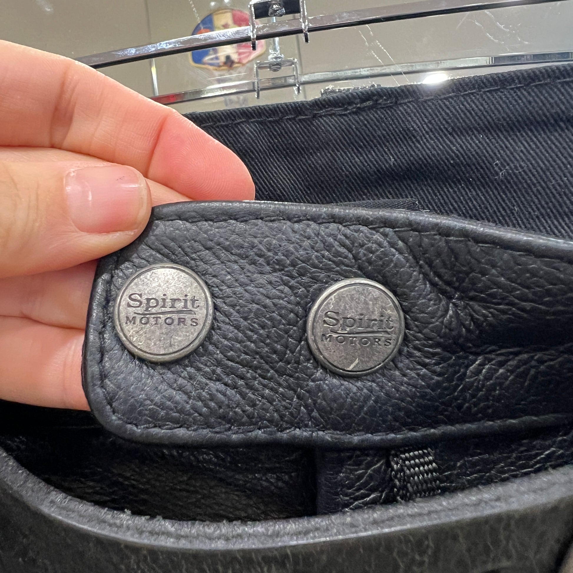 Close-up of a hand holding the inside flap of a black leather garment with two metal snap buttons engraved ‘Spirit Motors’