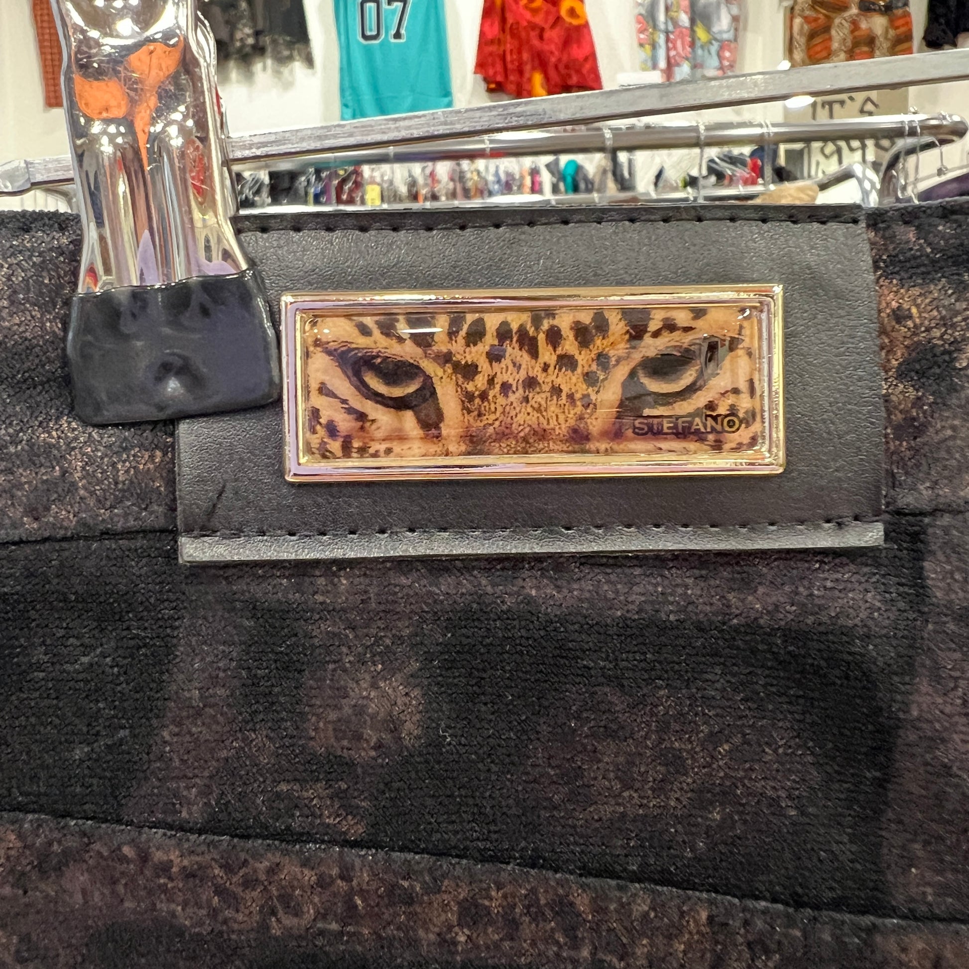 Close-up of a decorative plaque on a dark suede garment, featuring a gold-framed graphic of leopard eyes with the name “Stefano” on the right side
