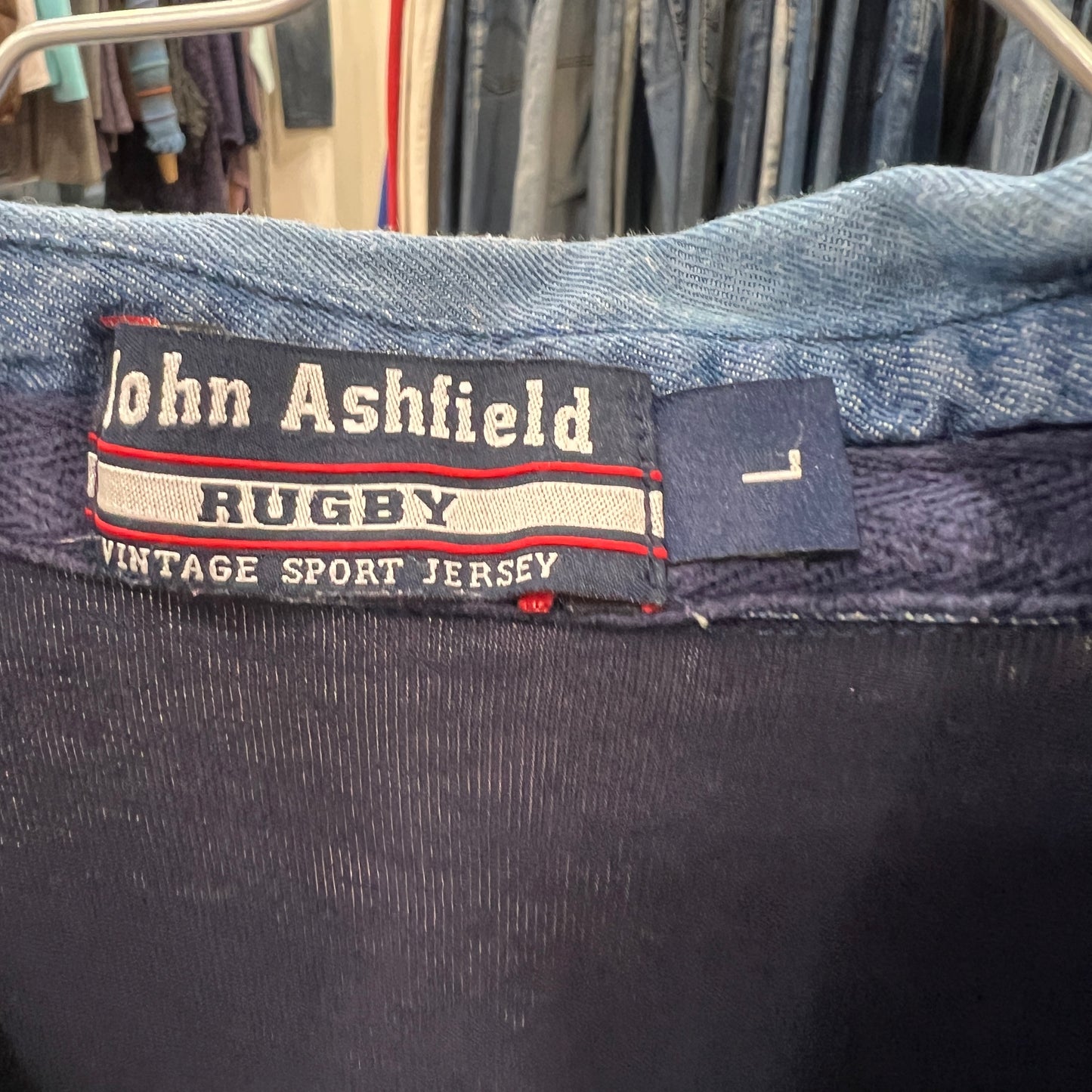 Close-up of a John Ashfield Rugby label on a garment with a clothing rack in the background