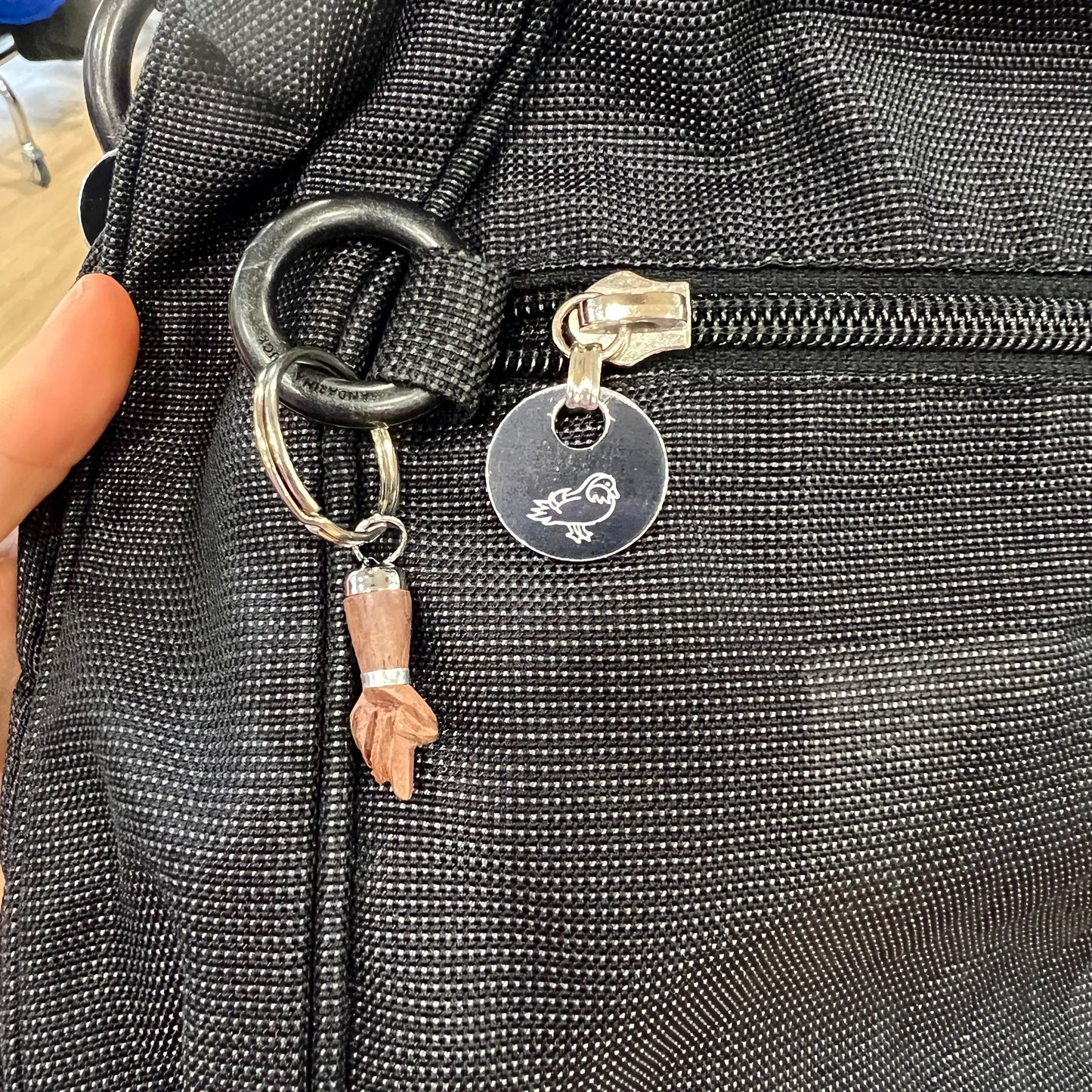 Close-up of a black textured bag with a zipper and two keychain charms attached: a round metal tag engraved with a small bird and a small carved wooden figurine hangin
