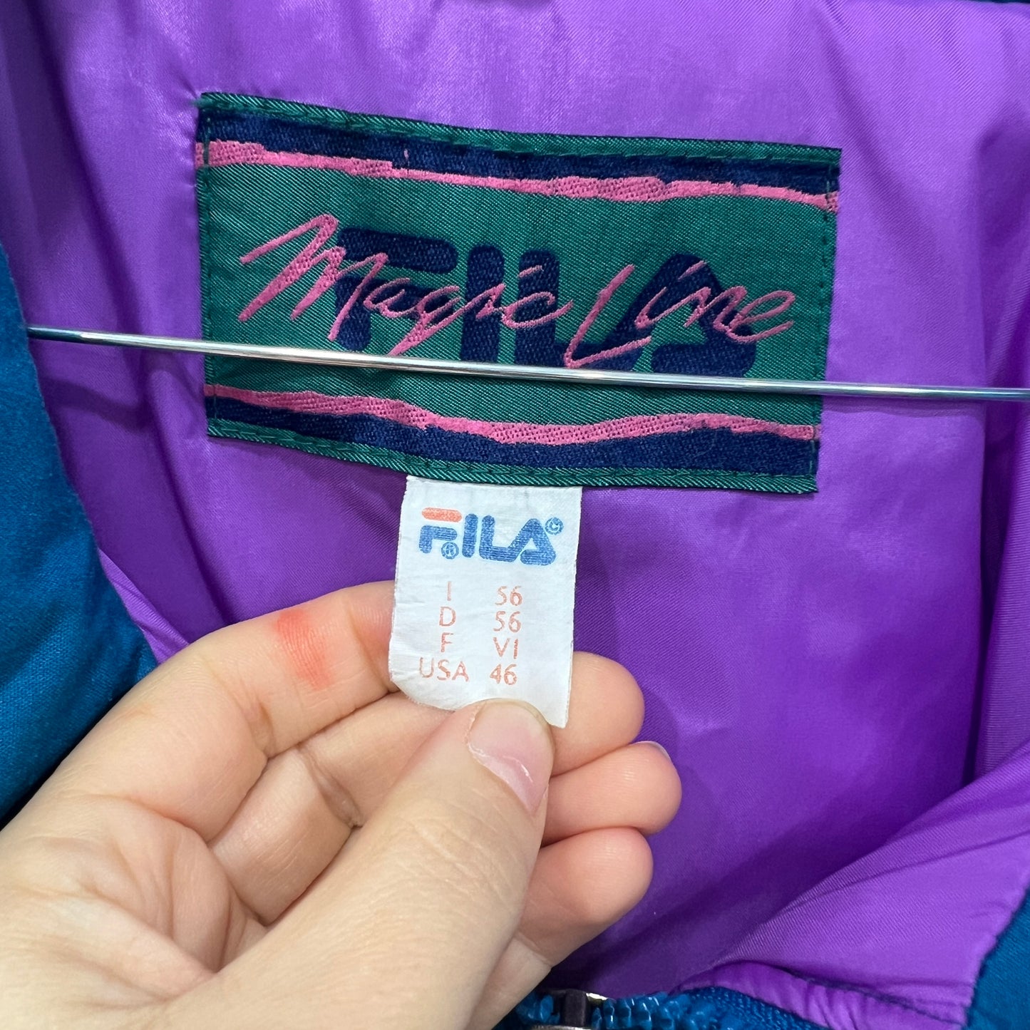 Close-up of a vintage FILA size tag held between two fingers, with a colorful “Magic Line” embroidered label on a purple jacket lining in the background
