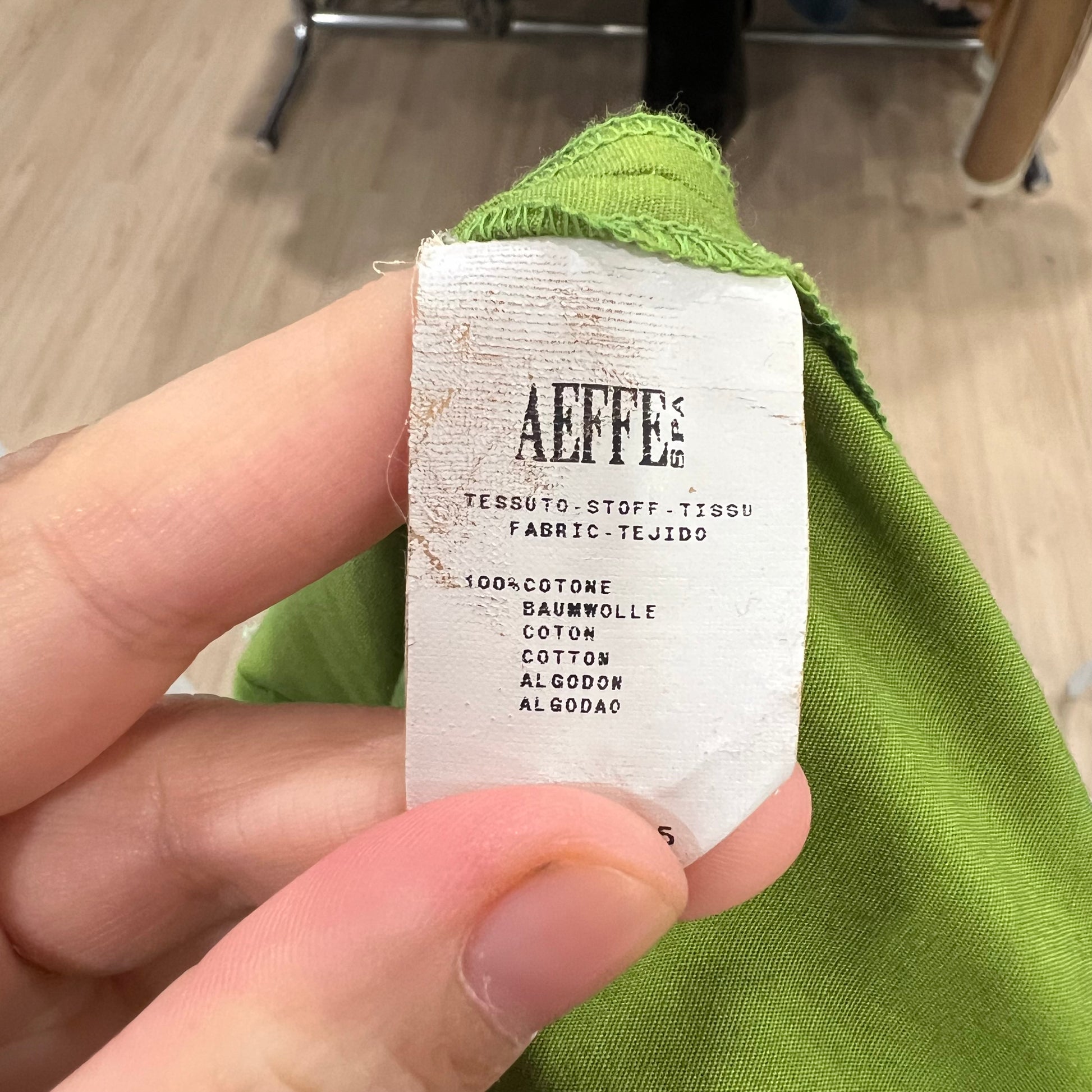 Hand holding a green fabric with an 'AEFFE' label on a wooden floor background