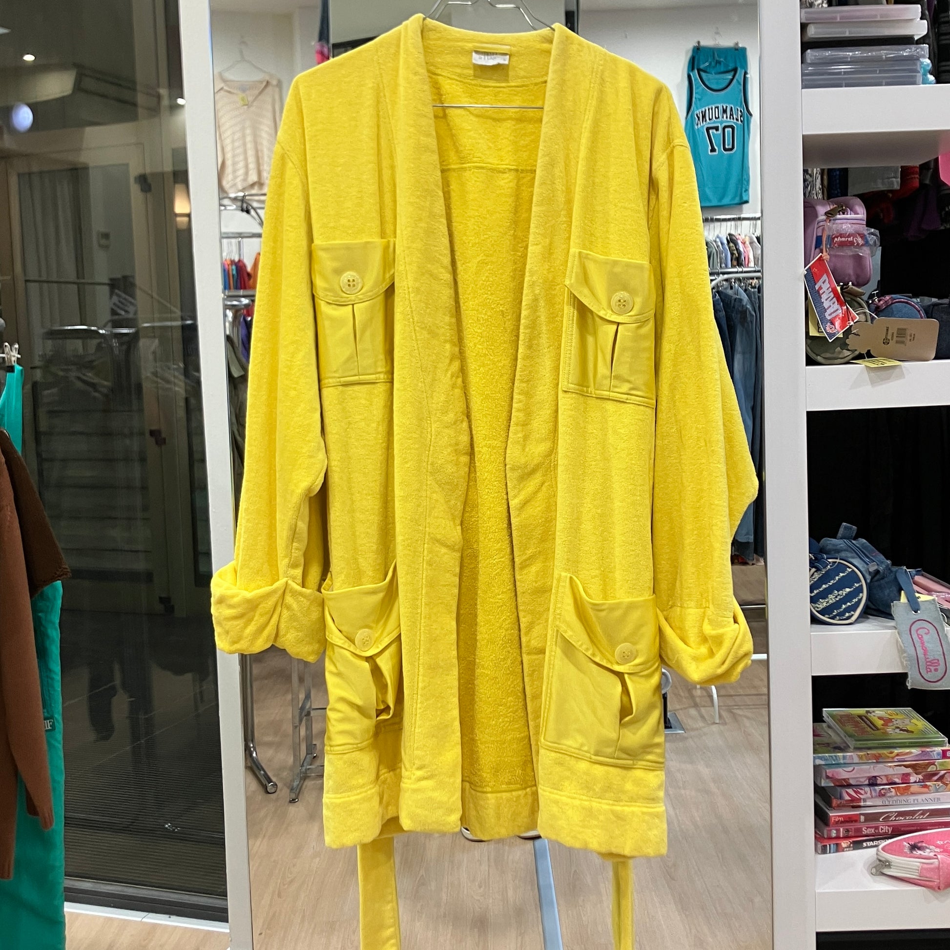 Front view of a bright yellow terry cloth bathrobe with multiple flap pockets, rolled sleeves, and a matching waist belt, displayed on a hanger inside a vintage clothing store
