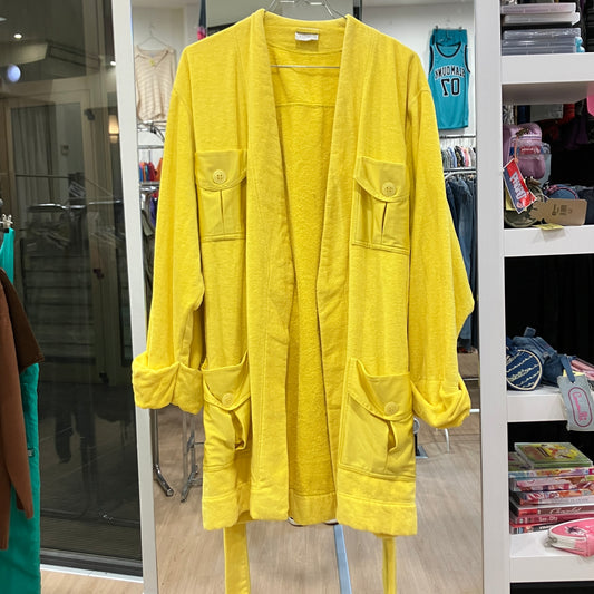 Front view of a bright yellow terry cloth bathrobe with multiple flap pockets, rolled sleeves, and a matching waist belt, displayed on a hanger inside a vintage clothing store
