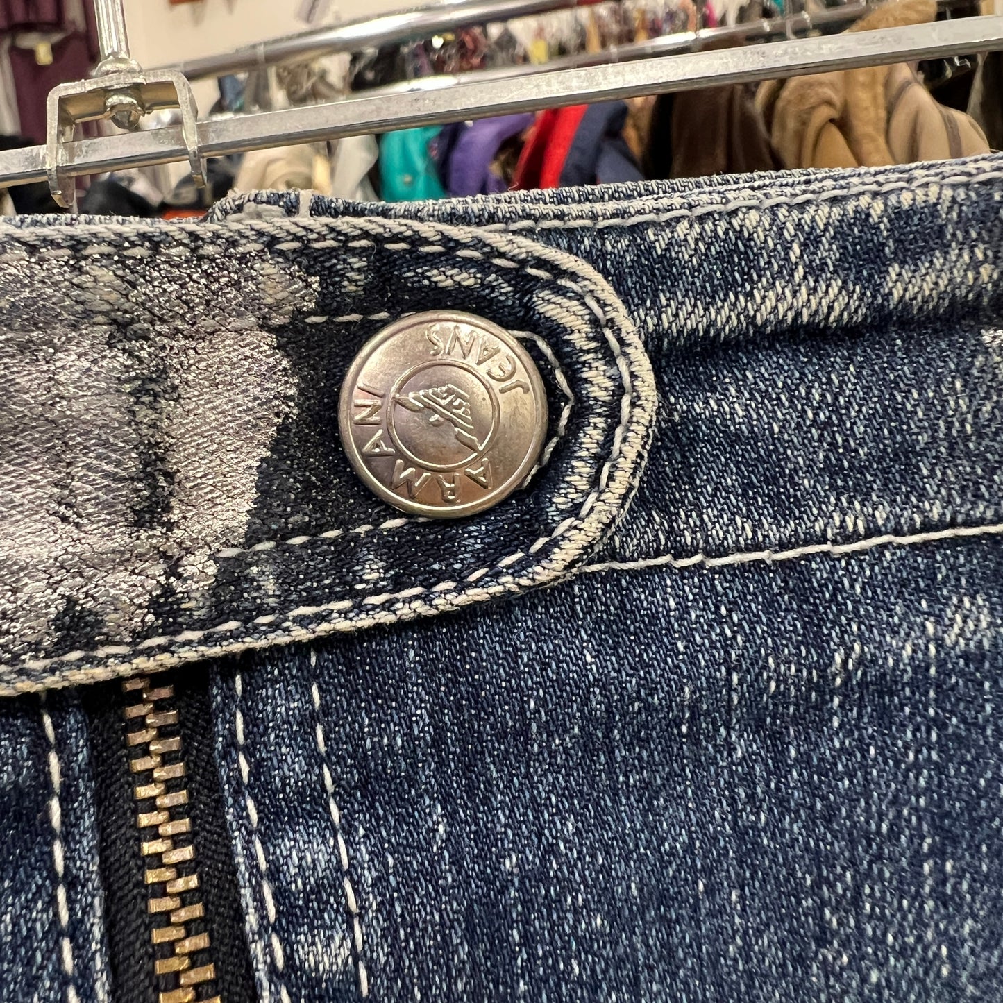 Close-up of the metal button on a pair of blue Armani Jeans denim pants, showing the engraved AJ logo and surrounding stitching with part of the zipper visible below
