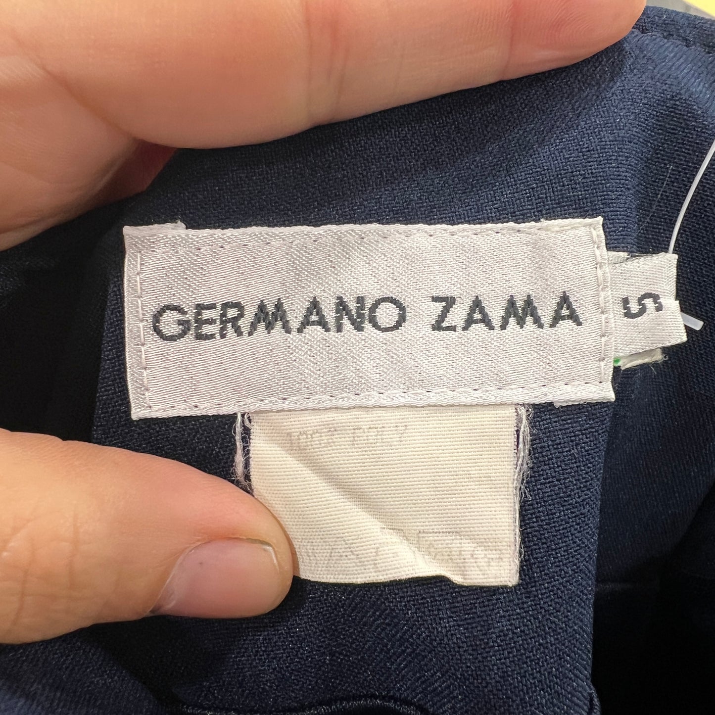 Close-up of a white Germano Zama brand label stitched onto a navy blue garment, held between two fingers
