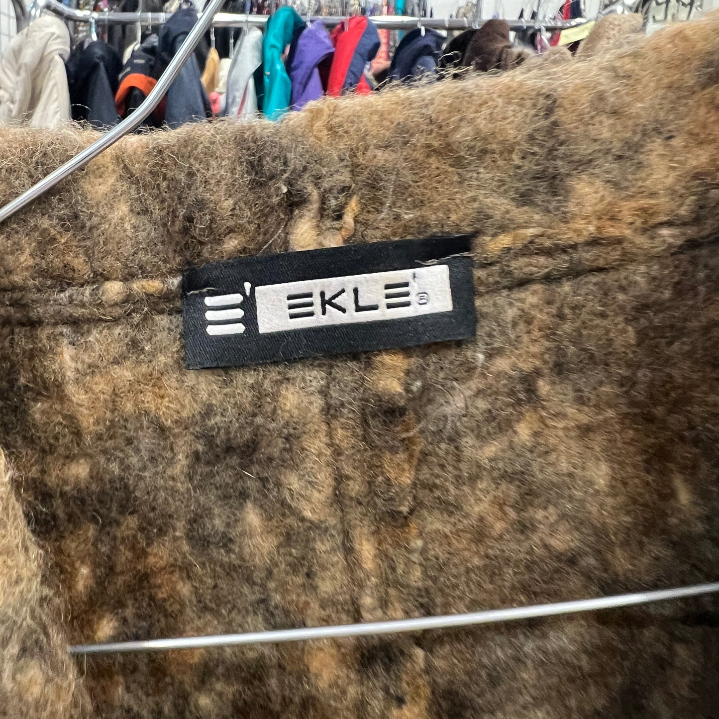 Close-up of a brown fur product with 'EKLE' label, background with clothes on hangers