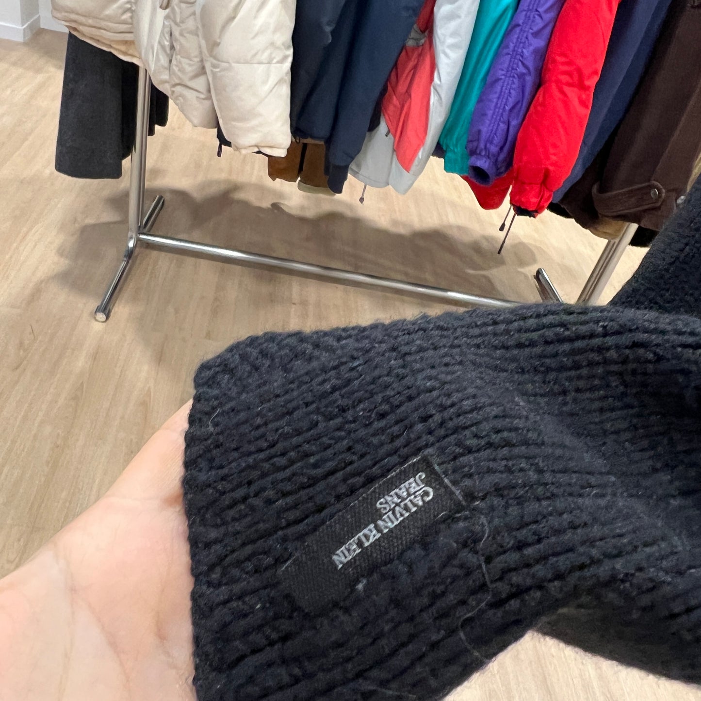 Black knit sweater with a brand label held in front of a rack of colorful coats