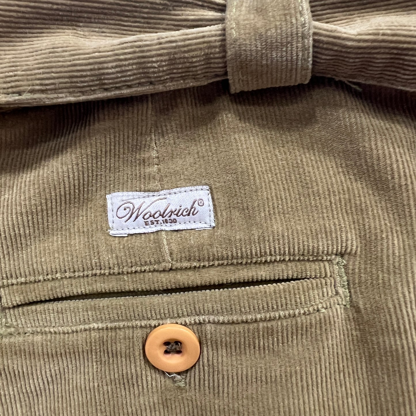 Close-up of tan corduroy trousers featuring a Woolrich label above a welt pocket with a beige button
