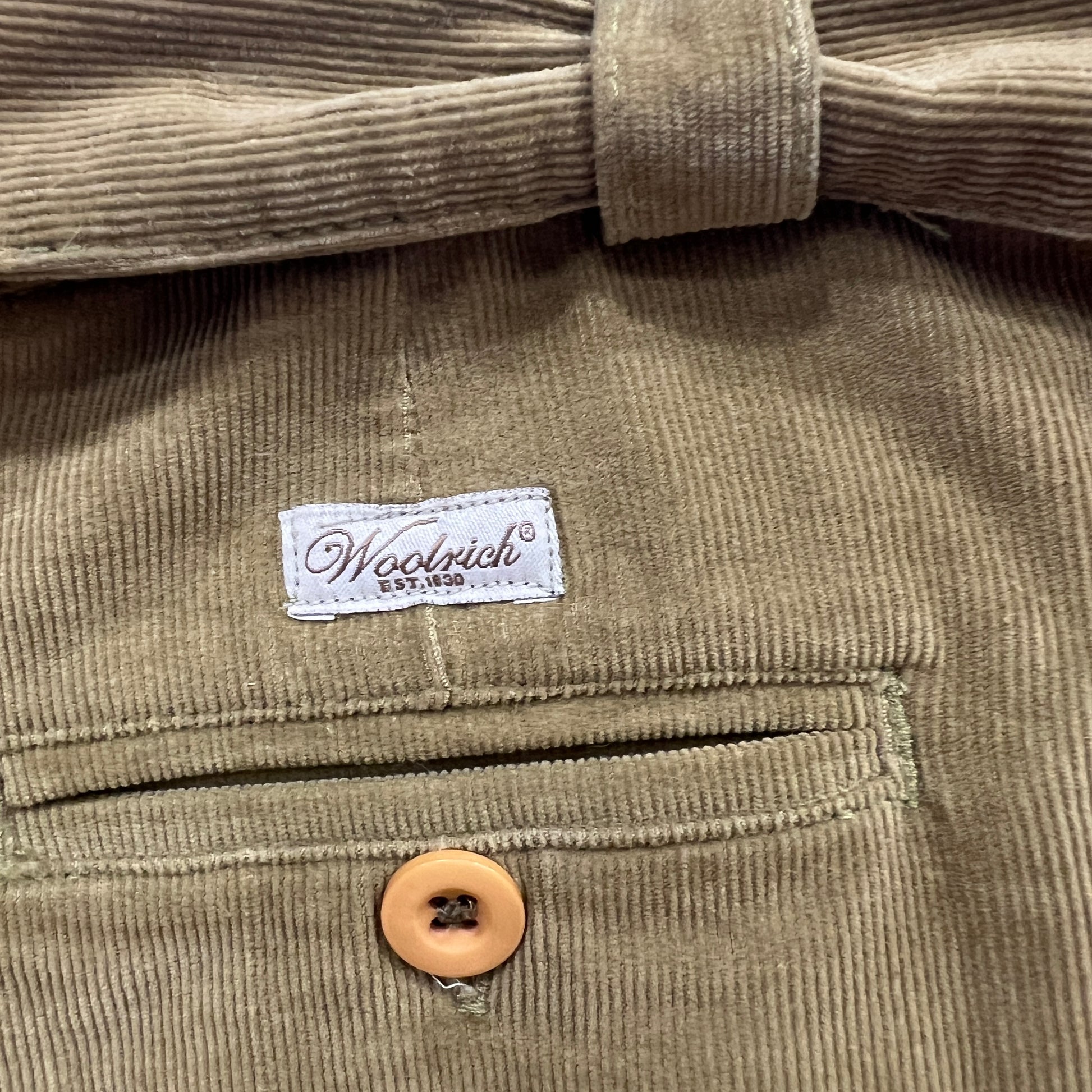 Close-up of tan corduroy trousers featuring a Woolrich label above a welt pocket with a beige button

