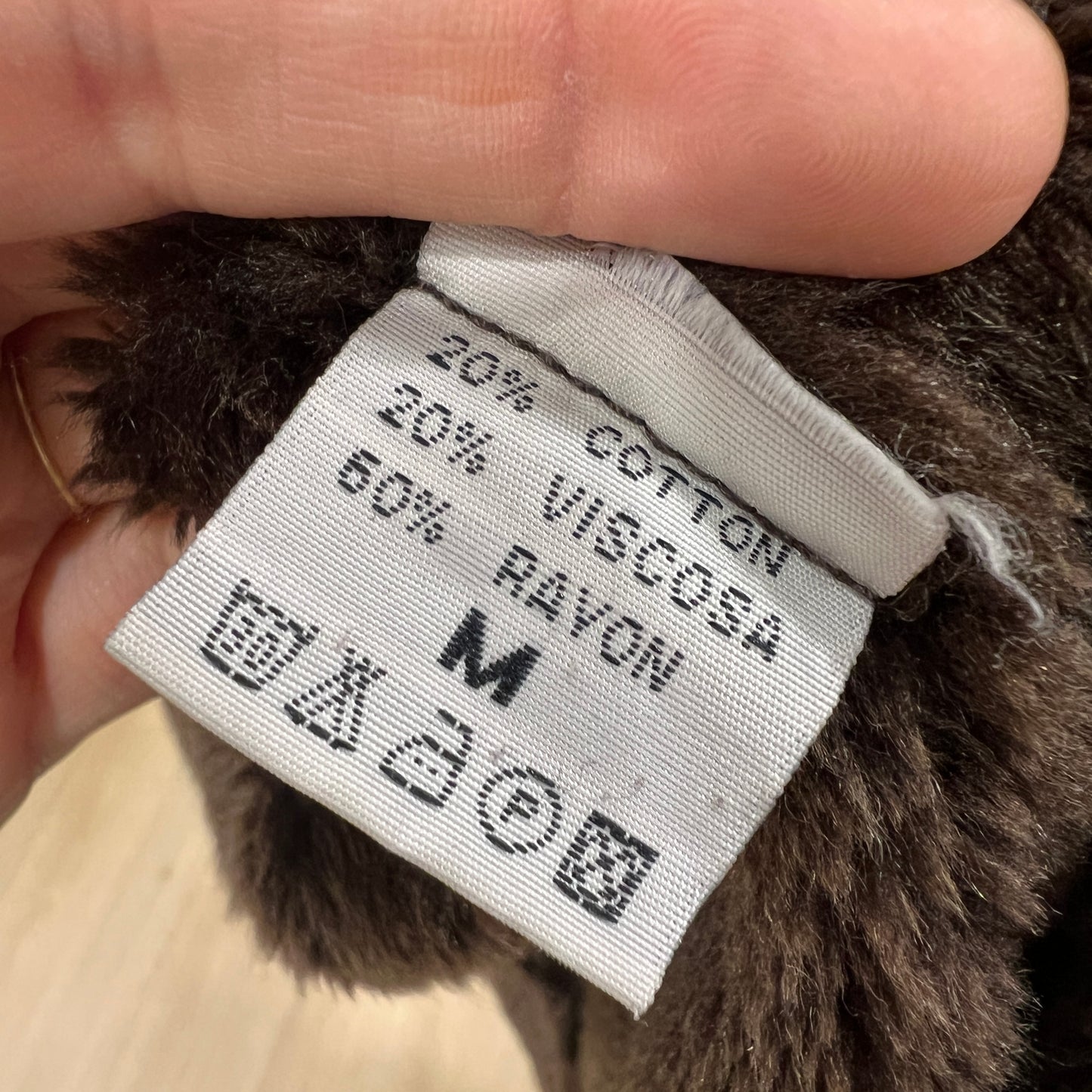 Close-up of an inner clothing label on a brown faux-fur garment showing composition 20% cotton, 20% viscose, 60% rayon and size M, held between two fingers