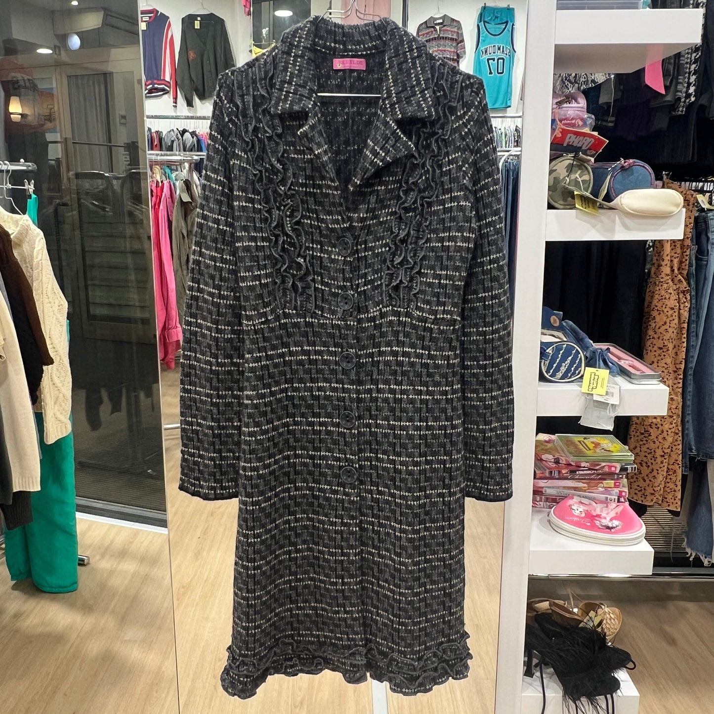 Full-length shot of a black and white textured long coat with ruffled details on the collar and hem, displayed on a hanger inside a vintage clothing store
