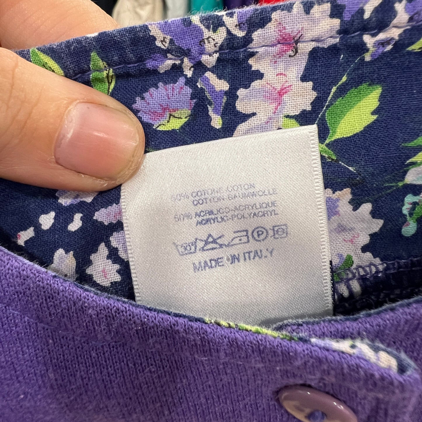Close-up of a white care and composition label on a floral-lined purple garment, showing fiber content, washing symbols, and “Made in Italy” while the tag is held between two fingers

