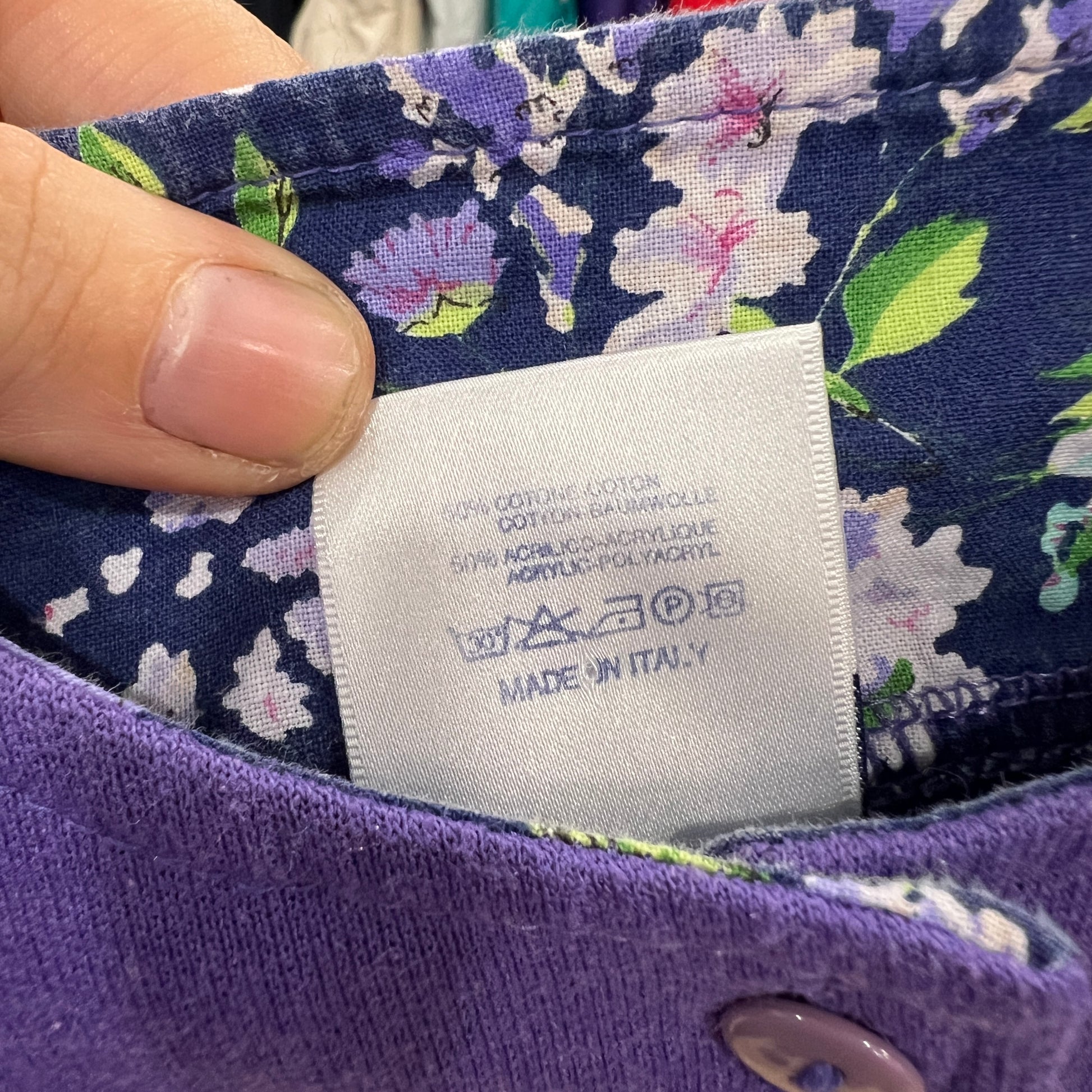 Close-up of a white care and composition label on a floral-lined purple garment, showing fiber content, washing symbols, and “Made in Italy” while the tag is held between two fingers
