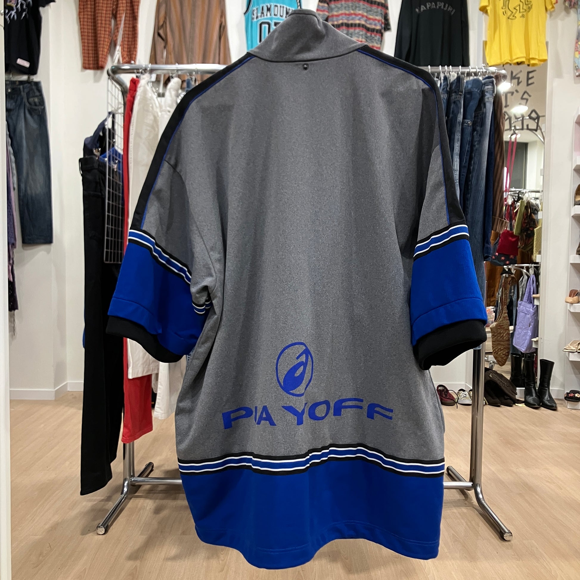 Back view of a vintage Asics oversized jacket in grey, black, and blue with bold blue branding and striped details, displayed on a hanger inside a vintage
