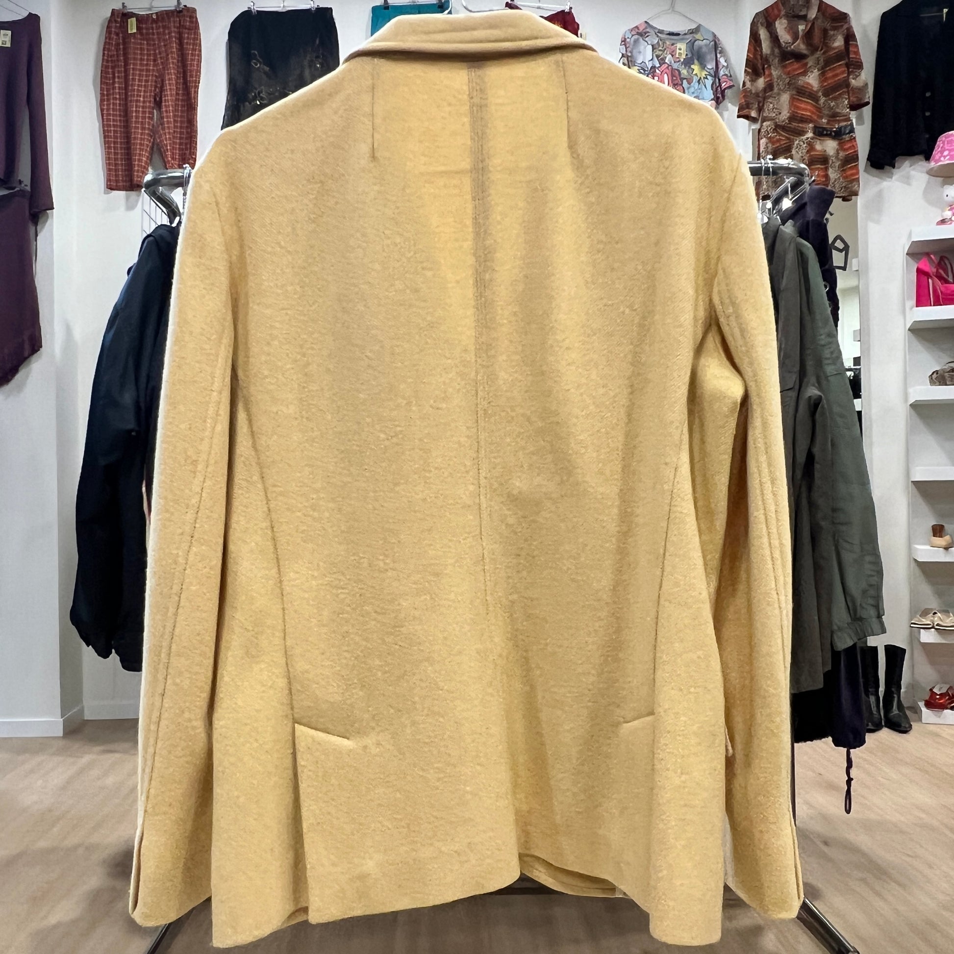 Back view of a beige wool blazer with a clean minimal design and subtle seam detailing, displayed on a hanger inside a vintage clothing store