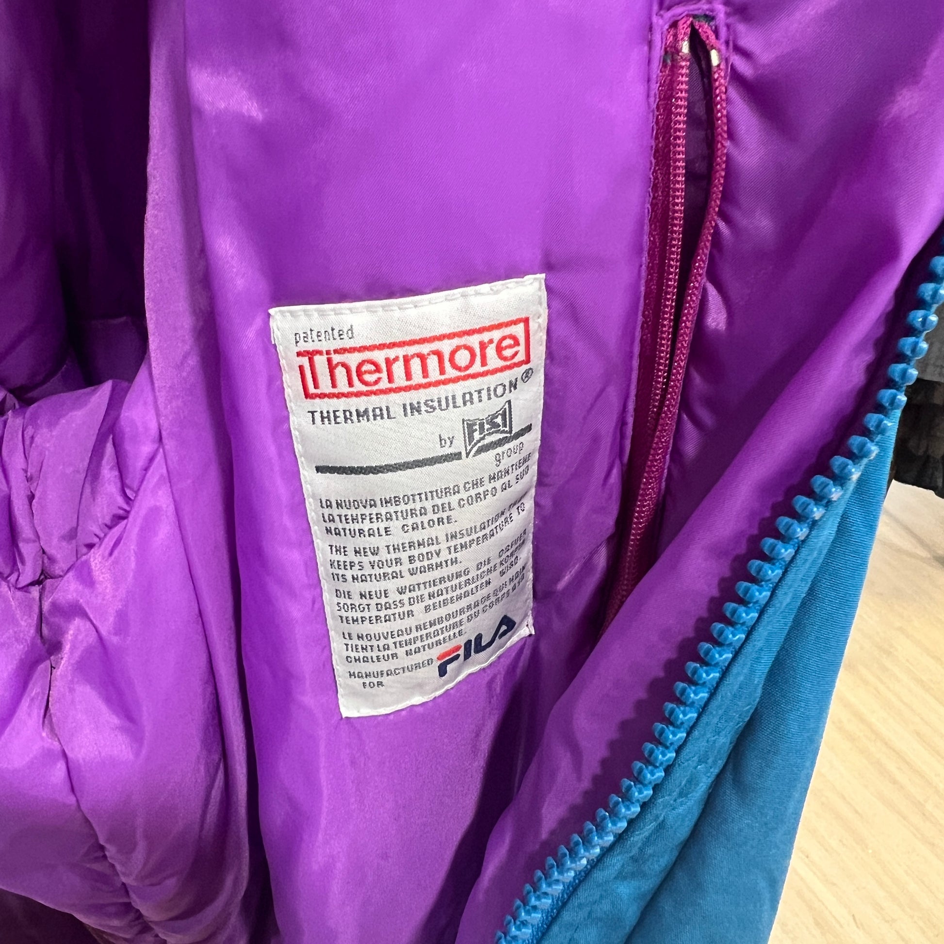 Close-up of a Thermore thermal insulation label stitched onto the purple lining of a vintage FILA jacket, with a blue zipper edge visible on the right
