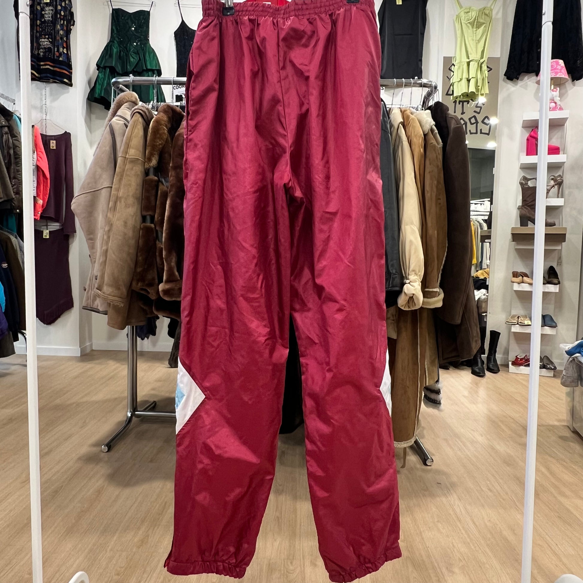Vintage red nylon track pants with elastic waist and white color block details on the legs hanging on a rack in a retro clothing store
