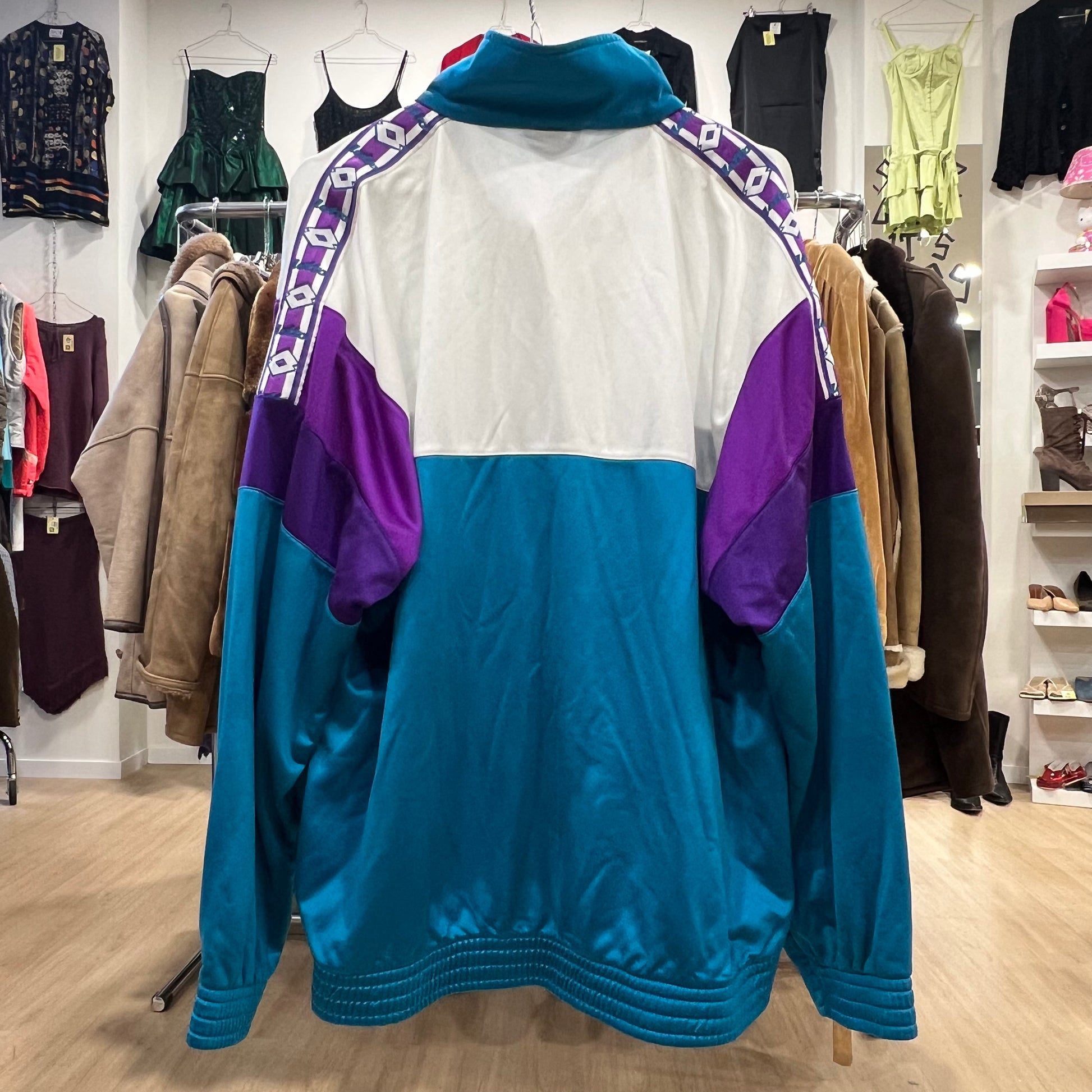 Vintage Lotto track jacket in teal, white, and purple color block design, featuring a zip-up front, high collar, and embroidered logo on the chest, displayed on a clothing rack in a store