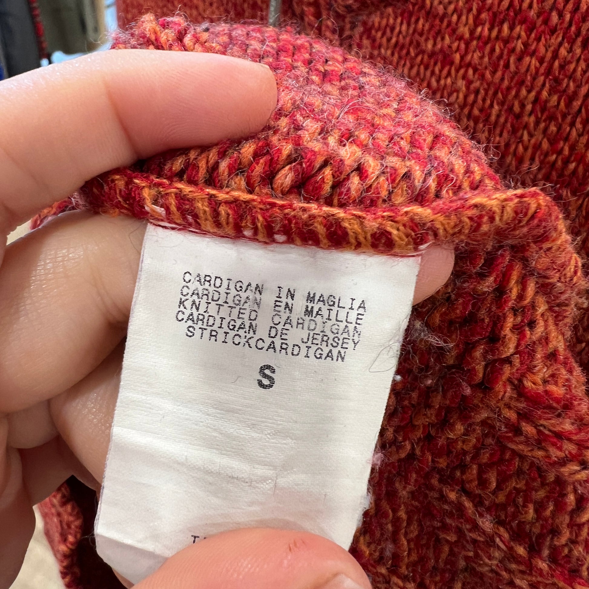 Close-up of a red knitted cardigan with a size label held by a hand