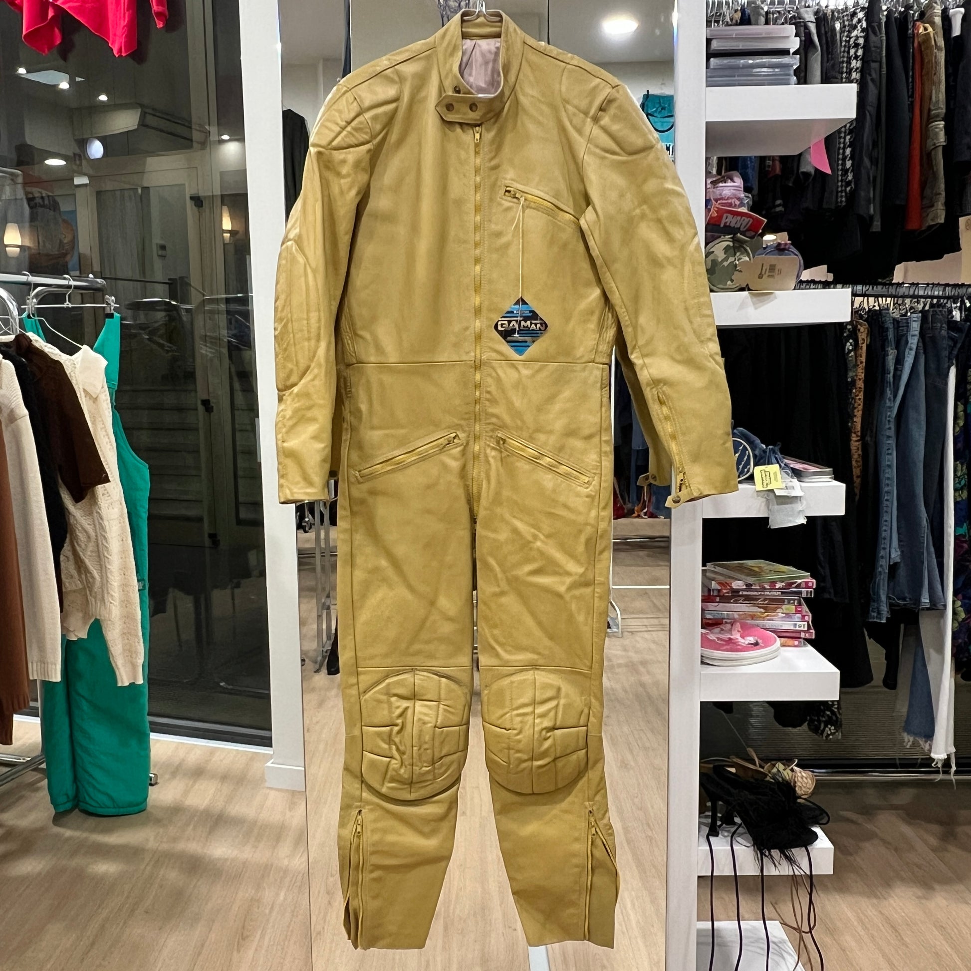 Full view of a yellow vintage work-style jumpsuit with zip pockets, padded knee panels, and a high collar, displayed on a hanger inside a vintage clothing store
