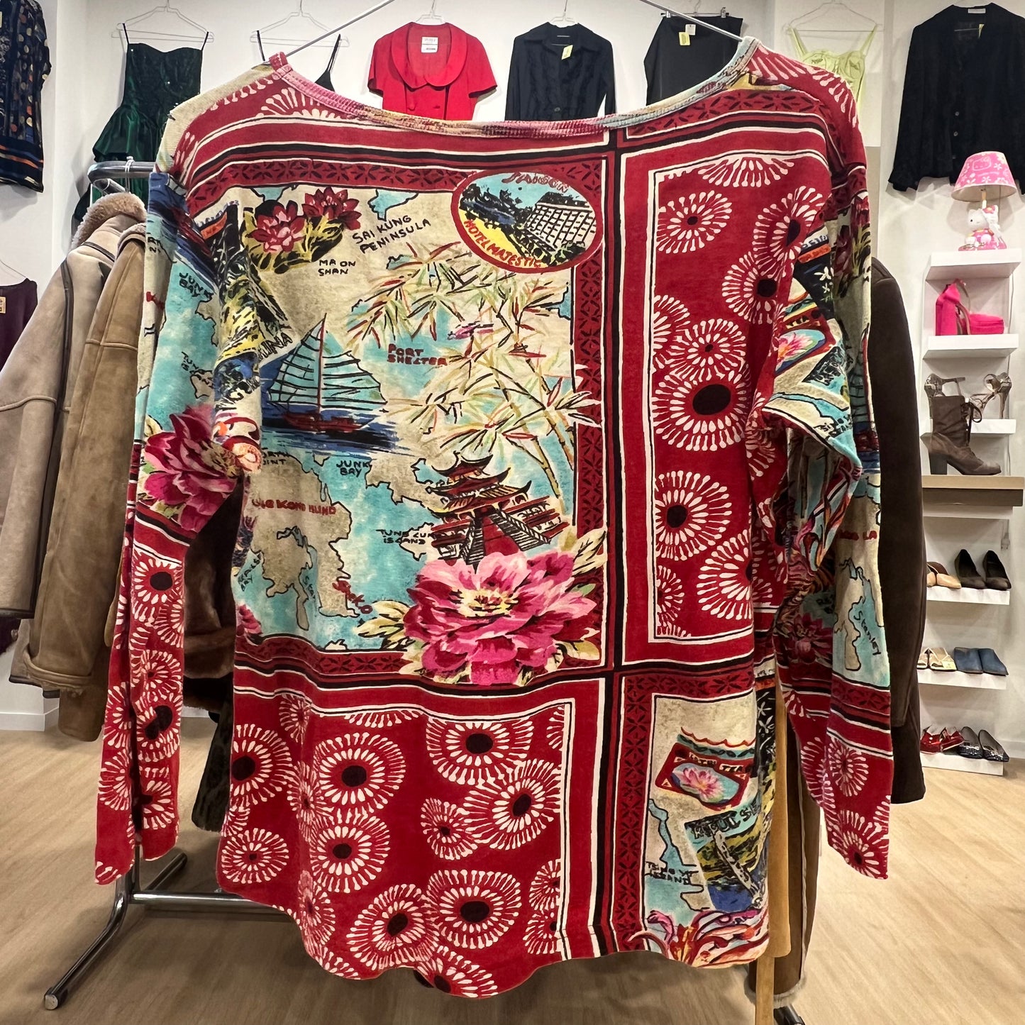 Long-sleeve bohemian top featuring a red base with a mixed travel and floral print, including maps, Asian motifs, and abstract circular patterns, displayed on a clothing rack in a store