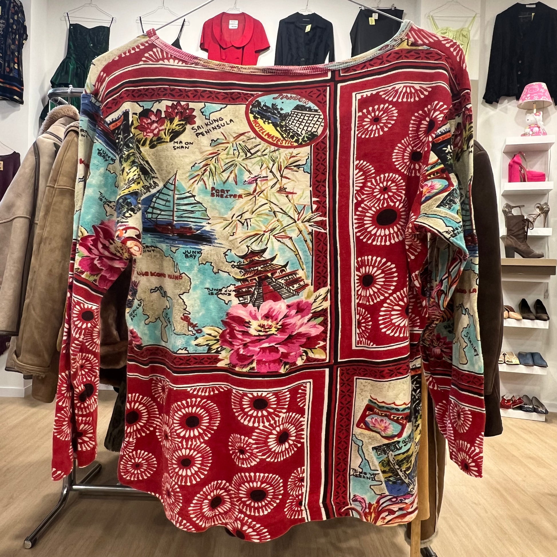 Long-sleeve bohemian top featuring a red base with a mixed travel and floral print, including maps, Asian motifs, and abstract circular patterns, displayed on a clothing rack in a store