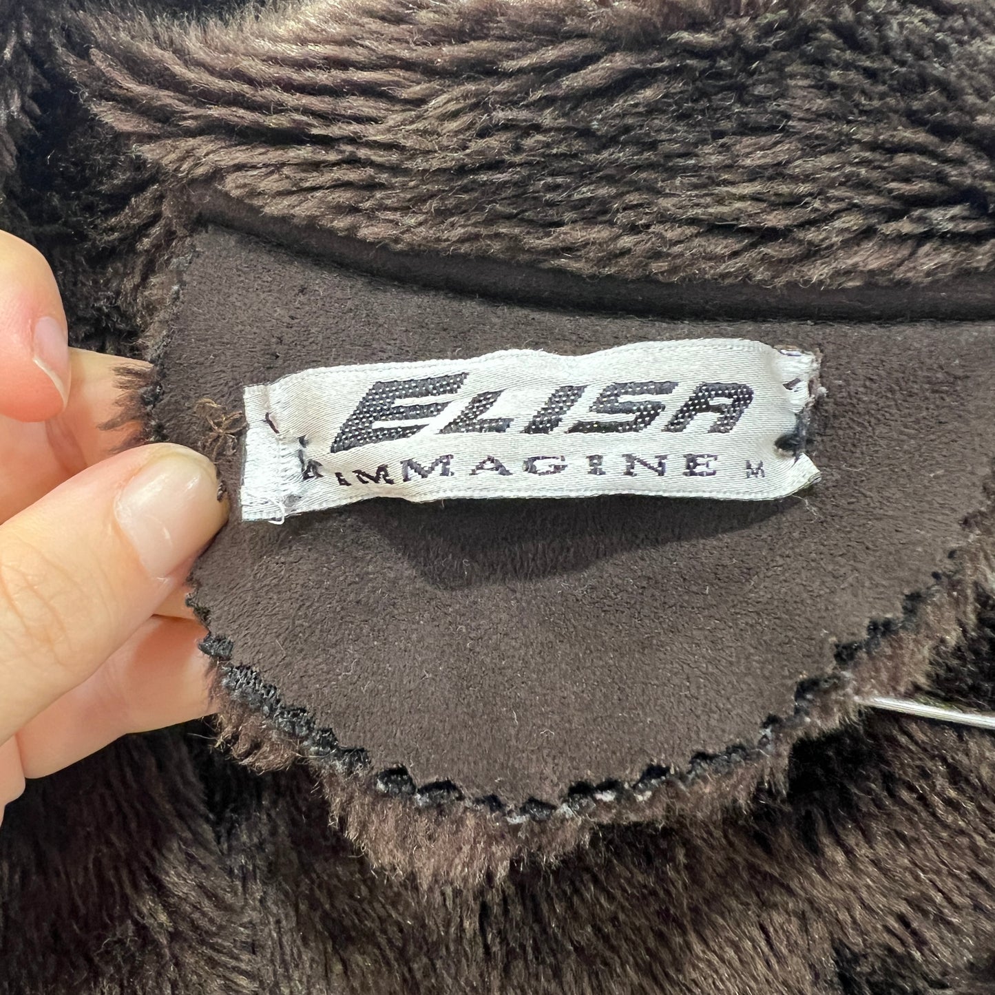 Close-up of a sewn-in brand label on a brown faux-fur garment, showing the text ‘ELISA IMMAGINE’ while the tag is held slightly lifted by two fingers