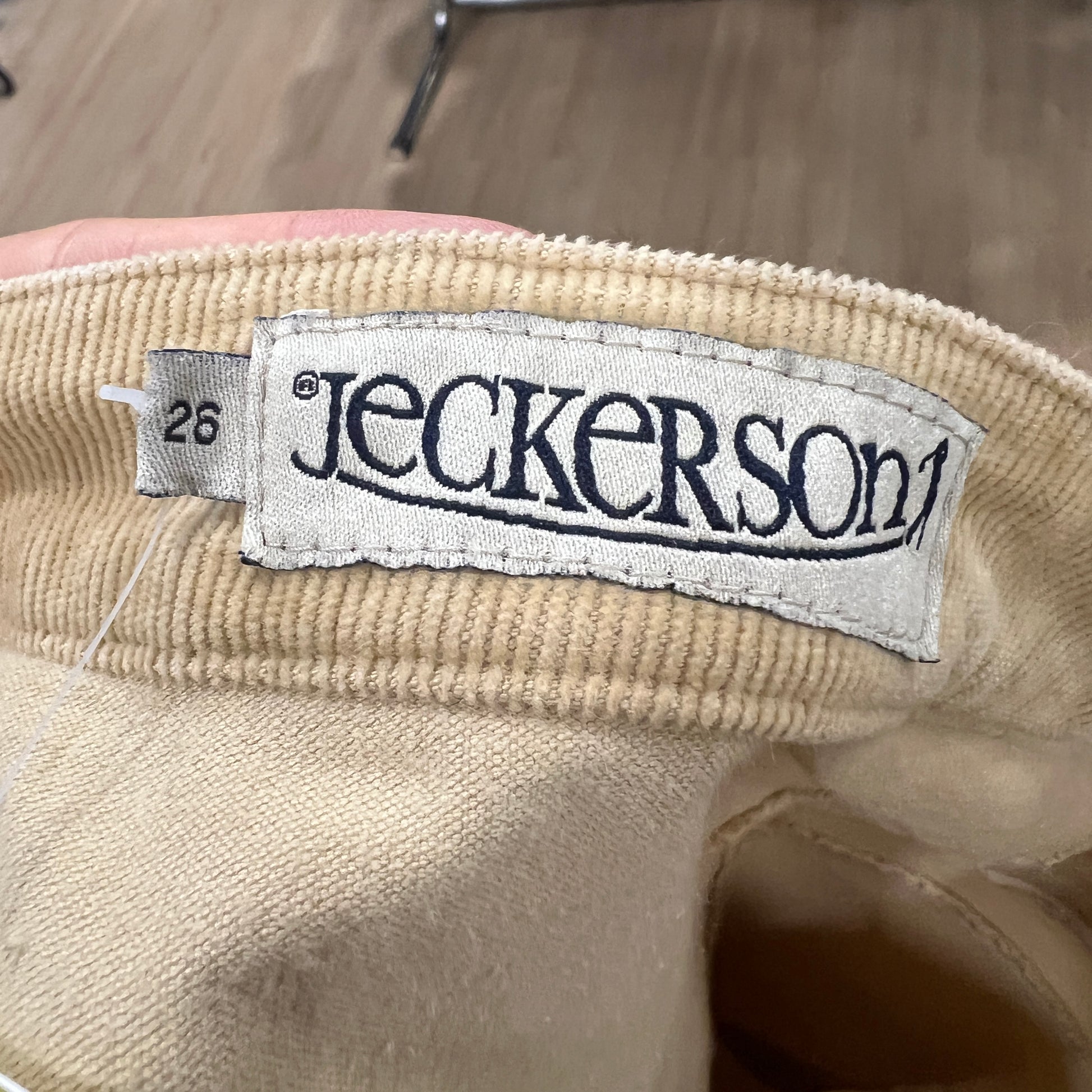 Close-up of a white Jeckerson label stitched onto the waistband of beige corduroy pants, with the size tag 26 visible on the left
