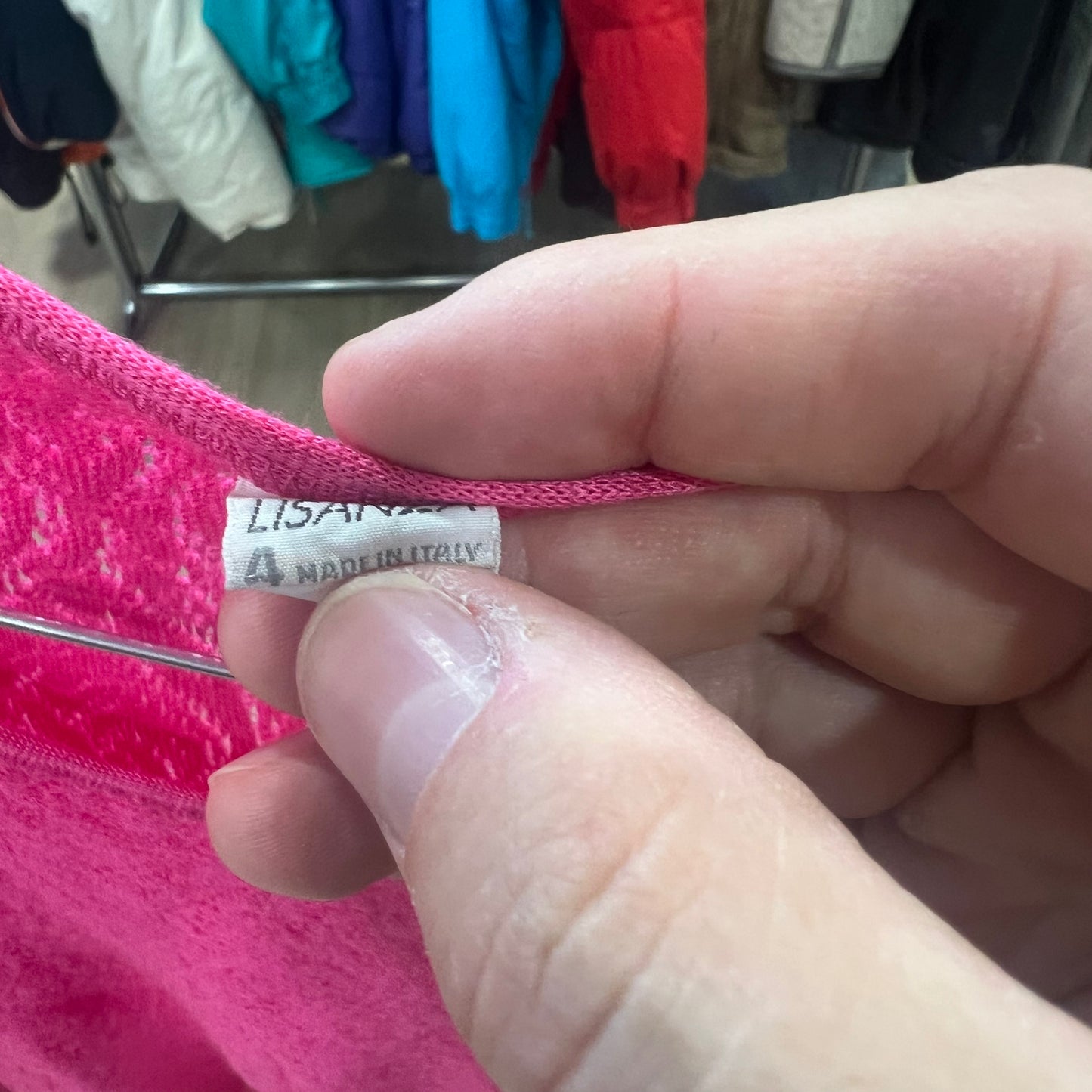 Close-up of a pink fabric with a label held by a hand, with a clothing rack in the background