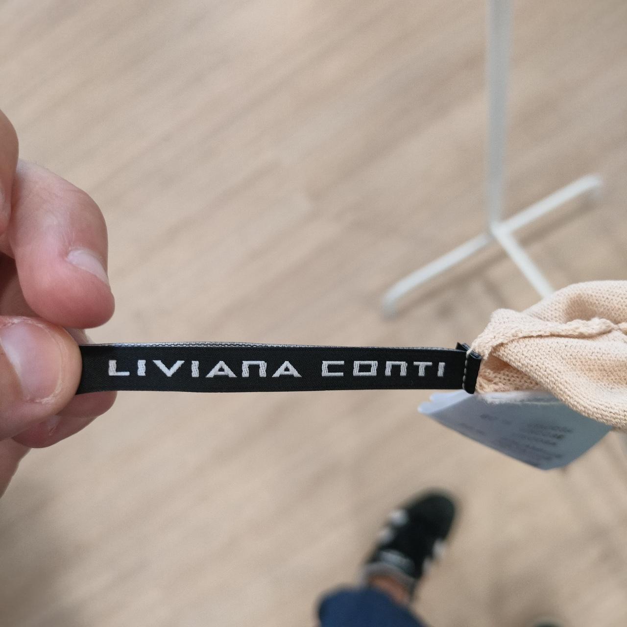 Person holding a black woven label with 'LIVIANA CONTI' text against a blurred background